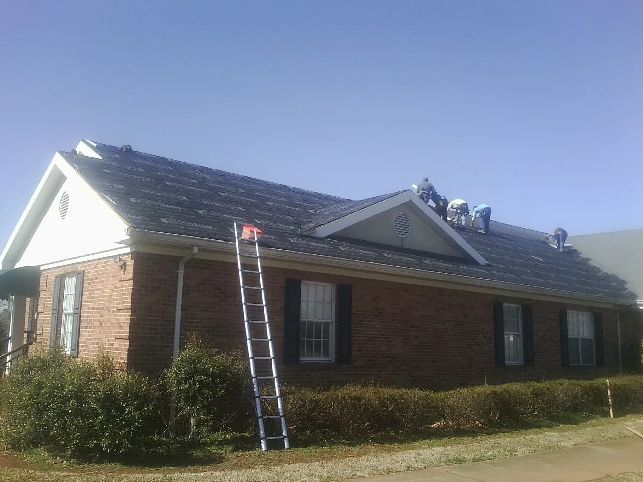 Crew tearing shingles off a brick ranch roof with a ladder set — Metal Roof Repair work by our crew in Sylacauga
