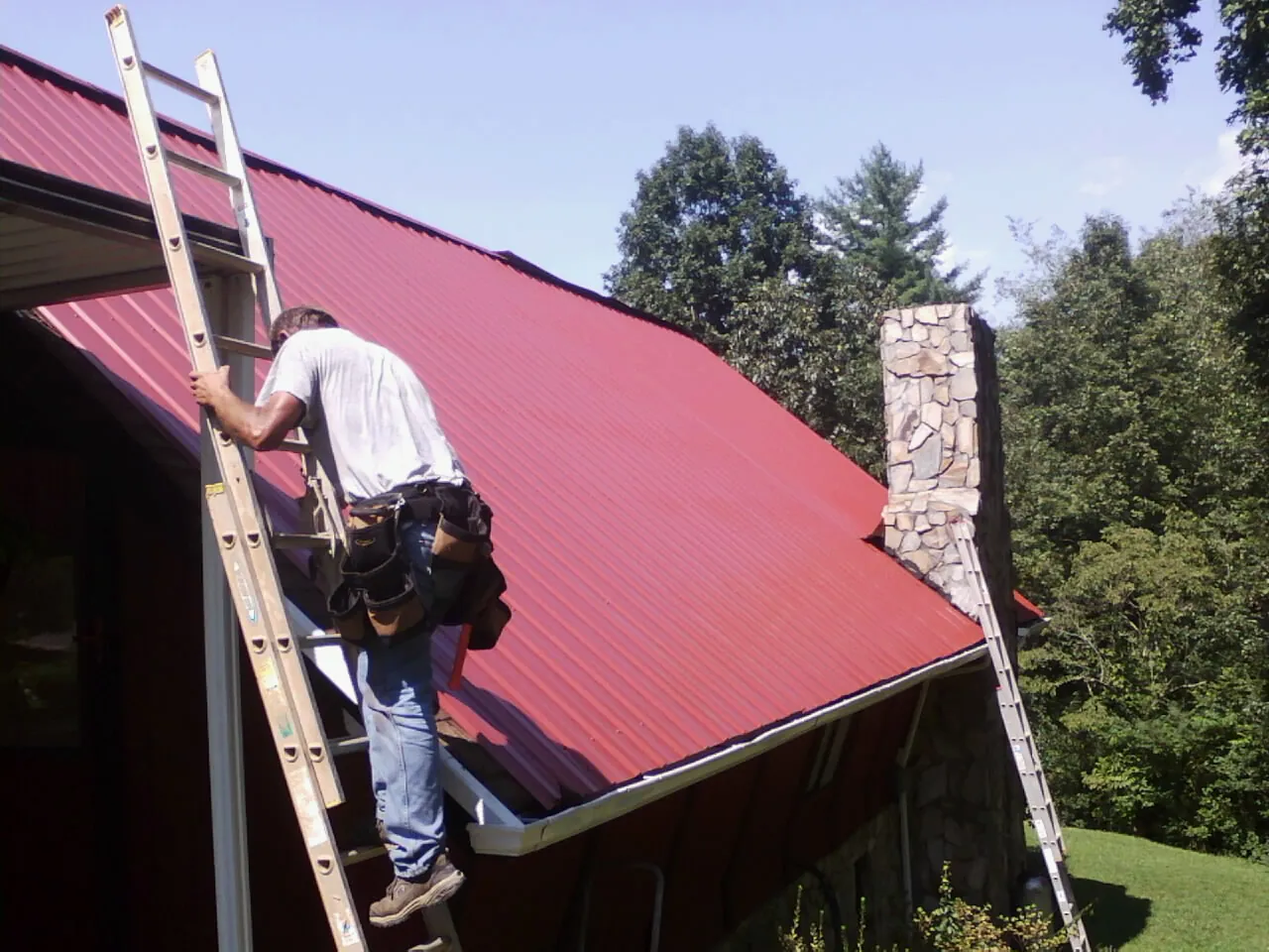 Roofer on a ladder beside a red metal roof and stone chimney — Soffit & Fascia Repair by Roofing Brothers in Sylacauga